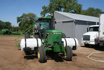 Green tractor equipped with two white saddle tanks for agricultural spraying, parked near a farm building.