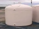 A beige cylindrical water storage tank with a domed lid, placed outdoors on a paved surface.