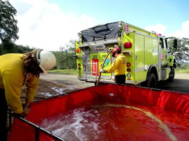 Red emergency water tank being filled by firefighters near a firetruck.