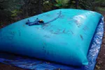 A blue water storage bladder placed on a tarp in an outdoor setting, designed for rainwater or other liquid storage.