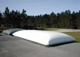 A long, rectangular white water bladder tank, placed on a paved outdoor area near a forest.