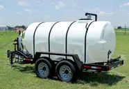 A white cylindrical trailer-mounted water tank with black straps, placed in an open grassy field.