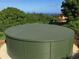 A round green corrugated steel water tank set in a lush green outdoor environment with trees and a house in the background.